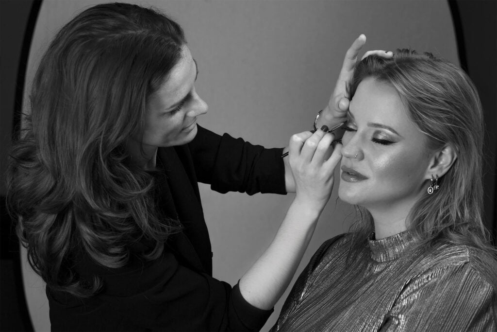 Studio photo of a working makeup artist finalizing the visage of her client/ model who is wearing a sparkling eyeshadow and bold eyeliner.