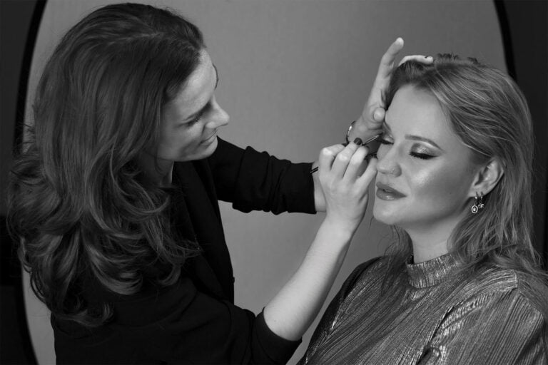 Studio photo of a working makeup artist finalizing the visage of her client/ model who is wearing a sparkling eyeshadow and bold eyeliner.